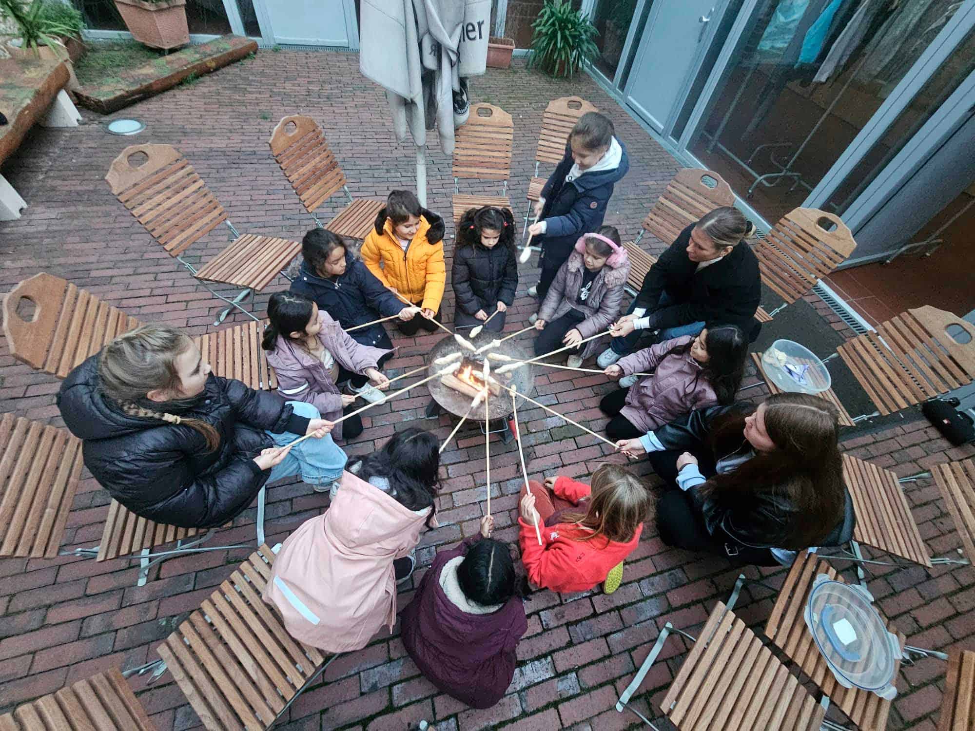 Kinder und Erwachsene sitzen im Kreis und zünden gemeinsam ein Lagerfeuer an.